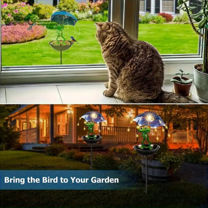 Cat looking out a window at a garden with a solar-powered bird feeder and bird on a stand.
